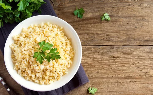 Eine weiße Schale ist gefüllt mit Bulgur aus Weizenkörnern. Dekoriert ist er mit frischer Petersilie. Neben der Schale liegen noch einzelne Blätter und Reste der Petersilie. Die Schale steht auf einem lilafarbenen Tischset, das auf einer dunklen, rustikalen Holzplatte liegt.