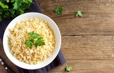 Eine weiße Schale ist gefüllt mit Bulgur aus Weizenkörnern. Dekoriert ist er mit frischer Petersilie. Neben der Schale liegen noch einzelne Blätter und Reste der Petersilie. Die Schale steht auf einem lilafarbenen Tischset, das auf einer dunklen, rustikalen Holzplatte liegt.