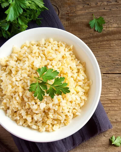 Eine weiße Schale ist gefüllt mit Bulgur aus Weizenkörnern. Dekoriert ist er mit frischer Petersilie. Neben der Schale liegen noch einzelne Blätter und Reste der Petersilie. Die Schale steht auf einem lilafarbenen Tischset, das auf einer dunklen, rustikalen Holzplatte liegt.