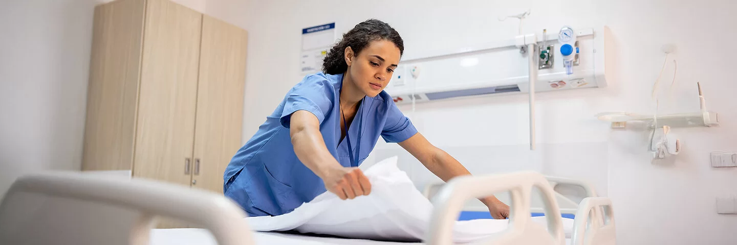 Eine junge Pflegefachfrau in blauem Kittel bereitet in einem Krankenhaus ein Patientenbett auf.