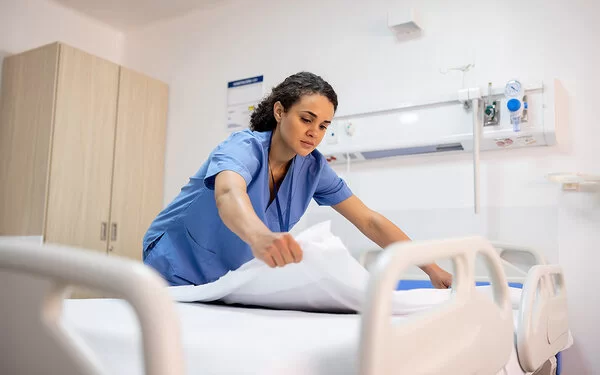 Eine junge Pflegefachfrau in blauem Kittel bereitet in einem Krankenhaus ein Patientenbett auf.