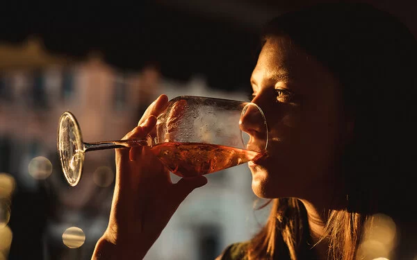 Eine Frau trinkt ein Glas Wein. Es ist Abend, das Licht ist gedämpft. Sie sitzt vermutlich in einer Bar.
