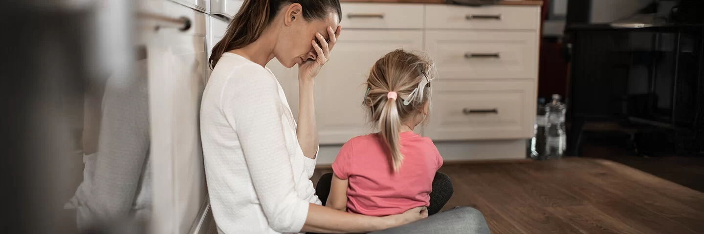 Eine Frau mit langen braunen Haaren sitzt im Schneidersitz in einer Küche auf dem Boden, an einen Schrank angelehnt. Sie wirkt erschöpft und hält sich eine Hand vor das Gesicht. Ein kleines Mädchen mit blondem Zopf sitzt auf ihrem Schoß, den Blick von den Betrachtern abgewandt.