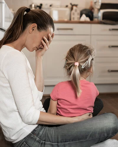 Eine Frau mit langen braunen Haaren sitzt im Schneidersitz in einer Küche auf dem Boden, an einen Schrank angelehnt. Sie wirkt erschöpft und hält sich eine Hand vor das Gesicht. Ein kleines Mädchen mit blondem Zopf sitzt auf ihrem Schoß, den Blick von den Betrachtern abgewandt.