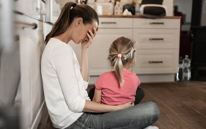 Eine Frau mit langen braunen Haaren sitzt im Schneidersitz in einer Küche auf dem Boden, an einen Schrank angelehnt. Sie wirkt erschöpft und hält sich eine Hand vor das Gesicht. Ein kleines Mädchen mit blondem Zopf sitzt auf ihrem Schoß, den Blick von den Betrachtern abgewandt.