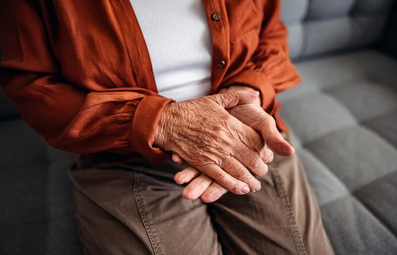 Nahaufnahme der faltigen Hände einer älteren Frau in braunen Hosen, weißem T-Shirt und einer orangefarbenen Bluse, die auf einem hellgrauen Sofa sitzt.