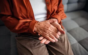 Nahaufnahme der faltigen Hände einer älteren Frau in braunen Hosen, weißem T-Shirt und einer orangefarbenen Bluse, die auf einem hellgrauen Sofa sitzt.