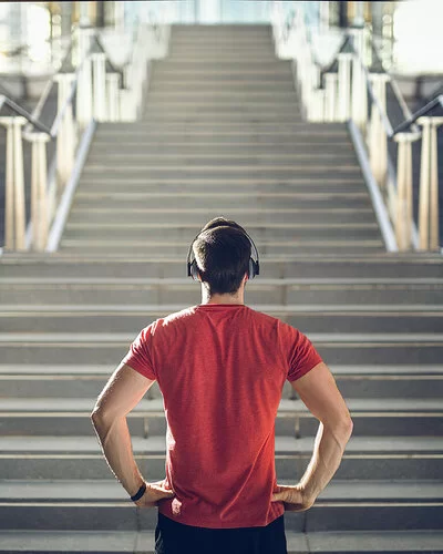 Ein Mann mit einem roten T-Shirt und Kopfhörern steht vor einer Treppe und blickt die Stufen herauf.