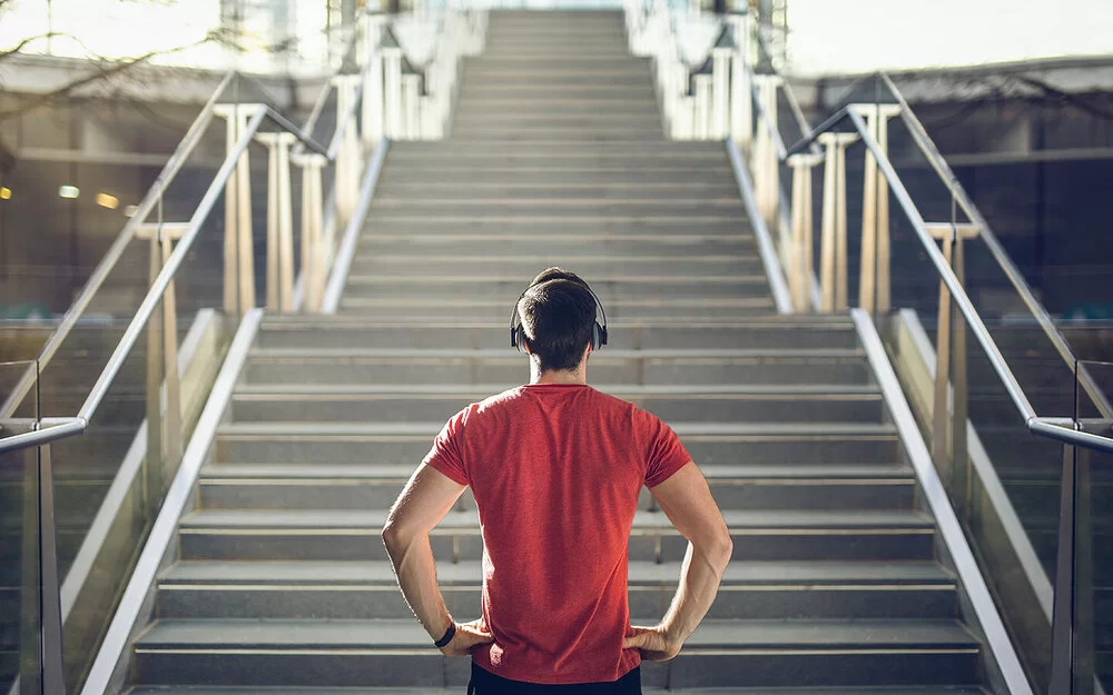 Ein Mann mit einem roten T-Shirt und Kopfhörern steht vor einer Treppe und blickt die Stufen herauf.