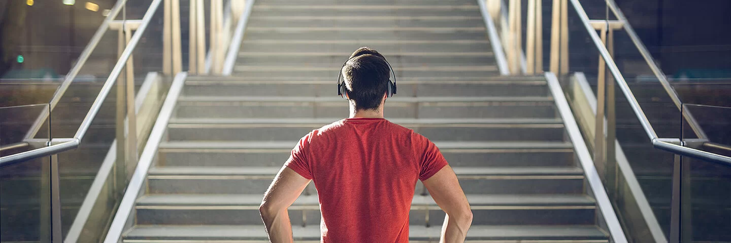 Ein Mann mit einem roten T-Shirt und Kopfhörern steht vor einer Treppe und blickt die Stufen herauf.