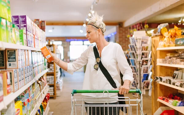 Eine Frau mit einem weißen Oberteil greift im Supermarktregal zu einer Packung Tee.