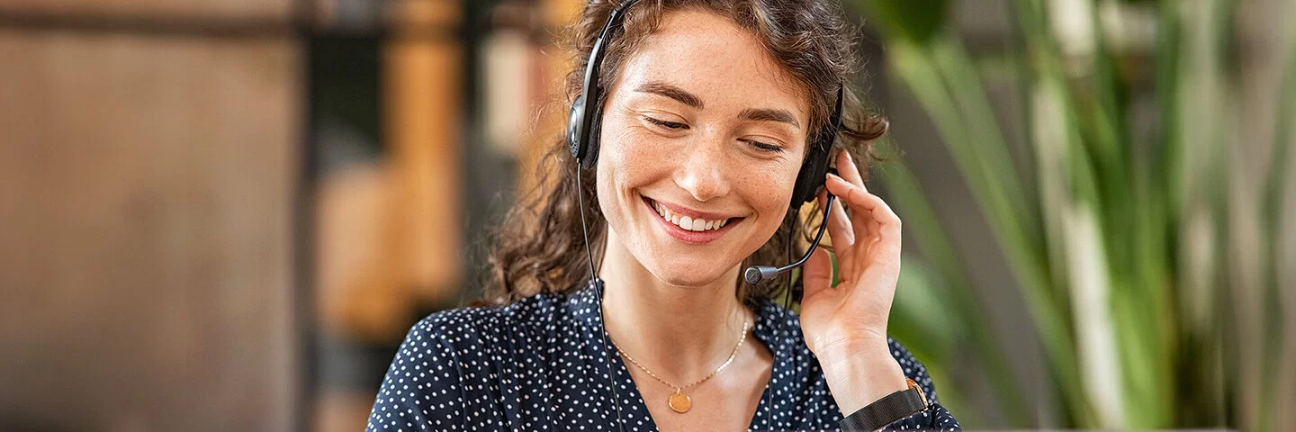 Eine junge Frau mit dunklem, lockigem Haar sitzt lächelnd mit einem Headset vor einem Laptop.