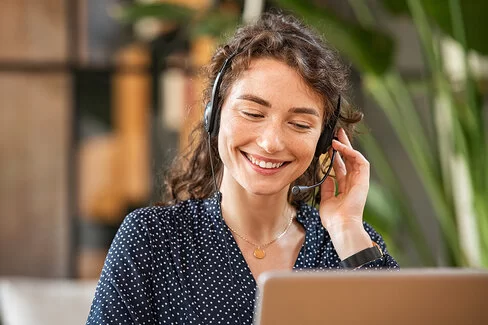 Eine junge Frau mit dunklem, lockigem Haar sitzt lächelnd mit einem Headset vor einem Laptop.