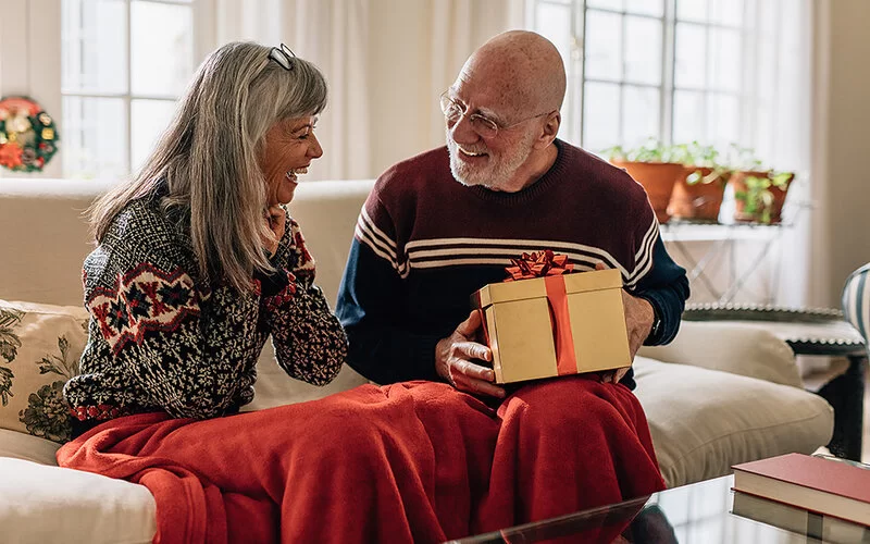 Ein älterer Mann und eine ältere Frau sitzen auf einem Sofa. Über ihren Beinen liegt eine kuschelige roteDecke. Der Mann hat ein in einer goldenen Schachtel verpacktes Geschenk auf den Knien und lacht die Frau an.