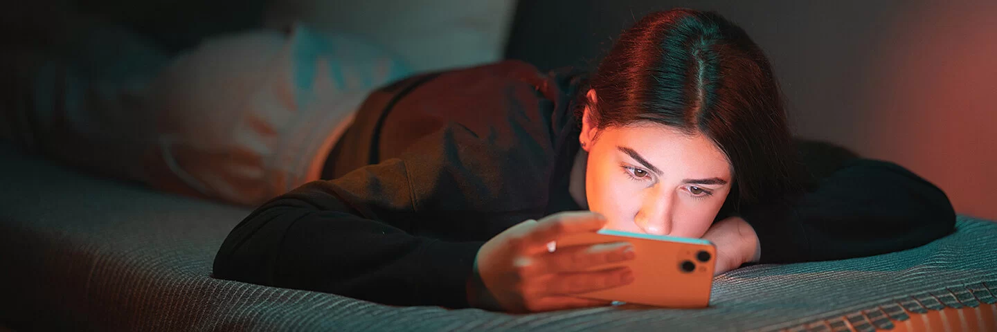 Ein junges Mädchen mit dunklen, in der Mitte gescheitelten Haaren bäuchlings auf dem Bett und schaut in ihr Handy. Das Zimmer ist dunkel, ihr Gesicht wird von dem Licht des Mobiltelefons erhellt.