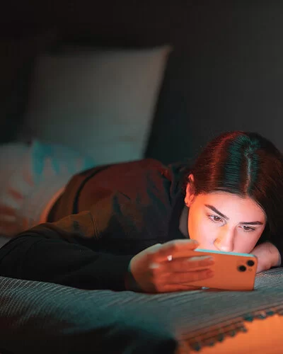 Ein junges Mädchen mit dunklen, in der Mitte gescheitelten Haaren bäuchlings auf dem Bett und schaut in ihr Handy. Das Zimmer ist dunkel, ihr Gesicht wird von dem Licht des Mobiltelefons erhellt.