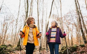 Zwei lachende Frauen mittleren Alters in Outdoor-Kleidung gehen mit Nordic-Walking-Stöcken durch einen herbstlichen Wald.