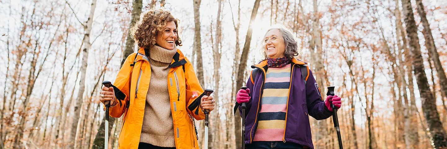 Zwei lachende Frauen mittleren Alters in Outdoor-Kleidung gehen mit Nordic-Walking-Stöcken durch einen herbstlichen Wald.