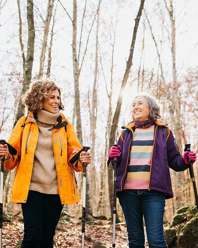 Zwei lachende Frauen mittleren Alters in Outdoor-Kleidung gehen mit Nordic-Walking-Stöcken durch einen herbstlichen Wald.