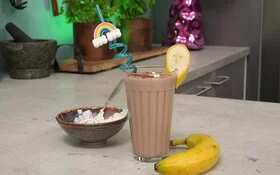 Schoko-Bananen-Proteinshake nach einem Rezept von Felicitas Then, angerichtet in einem hohen Glas mit buntem Strohhalm und Bananenscheibe am Rand. Rechts liegt eine Banane, links steht eine braune Schale mit Magerquark.