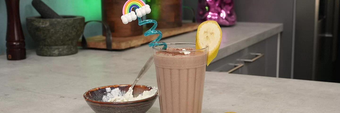 Schoko-Bananen-Proteinshake nach einem Rezept von Felicitas Then, angerichtet in einem hohen Glas mit buntem Strohhalm und Bananenscheibe am Rand. Rechts liegt eine Banane, links steht eine braune Schale mit Magerquark.