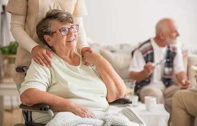 Eine ältere Frau sitzt im Rollstuhl. Sie wirkt zufrieden und hält die Hand einer Pflegerin, die hinter ihr steht. Im Hintergrund sitzt ein älterer Mann auf einer Couch und unterhält sich mit einer weiteren anwesenden Person.