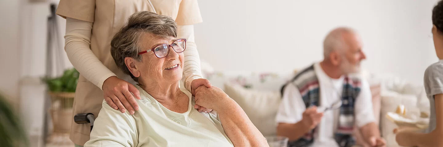 Eine ältere Frau sitzt im Rollstuhl. Sie wirkt zufrieden und hält die Hand einer Pflegerin, die hinter ihr steht. Im Hintergrund sitzt ein älterer Mann auf einer Couch und unterhält sich mit einer weiteren anwesenden Person.