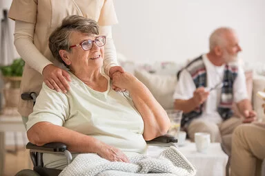 Eine ältere Frau sitzt im Rollstuhl. Sie wirkt zufrieden und hält die Hand einer Pflegerin, die hinter ihr steht. Im Hintergrund sitzt ein älterer Mann auf einer Couch und unterhält sich mit einer weiteren anwesenden Person.