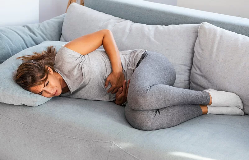 Eine junge Frau in grauen Leggings und hellgrauem T-Shirt liegt auf einem grauen Sofa und hält sich mit beiden Händen den Bauch. Sie hat offensichtlich dort Schmerzen.