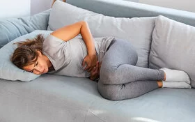 Eine junge Frau in grauen Leggings und hellgrauem T-Shirt liegt auf einem grauen Sofa und hält sich mit beiden Händen den Bauch. Sie hat offensichtlich dort Schmerzen.
