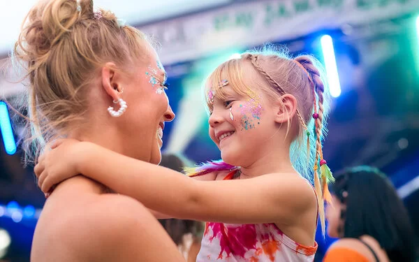 Eine Mutter hat auf einem Musikfestival im Sommer ihre Tochter auf dem Arm, beide mit Glitzer im Gesicht und sie lächeln sich an. Im Hintergrund sind die Lichter einer Musikbühne zu sehen.