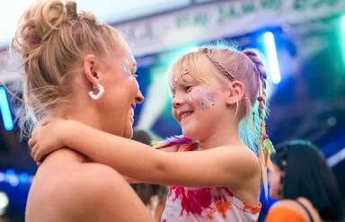 Eine Mutter hat auf einem Musikfestival im Sommer ihre Tochter auf dem Arm, beide mit Glitzer im Gesicht und sie lächeln sich an. Im Hintergrund sind die Lichter einer Musikbühne zu sehen.