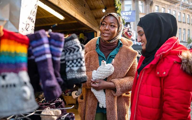 Zwei junge Frauen mit Kopftuch stehen auf einem Weihnachtsmarkt vor einem Verkaufsstand und probieren Handschuhe an.