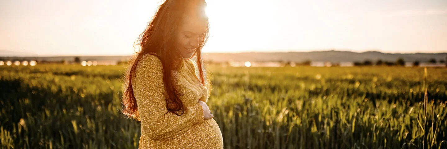 Eine junge, schwangere Frau steht in einem Feld und hält sich lächelnd den Bauch.