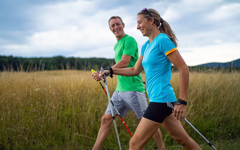 Ein Paar mittleren Alters betreibt gemeinsam Nordic Walking und lacht miteinander. Im Hintergrund ist ein Feld zu sehen.