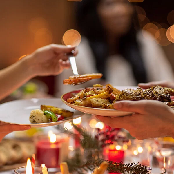 In einer Nahaufnahme ist eine festlich gedeckte Weihnachtstafel zu sehen. Kerzen brennen, im Ofen gebackene Gemüsestücke werden auf einen Teller gegeben.