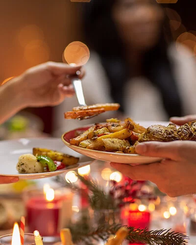 In einer Nahaufnahme ist eine festlich gedeckte Weihnachtstafel zu sehen. Kerzen brennen, im Ofen gebackene Gemüsestücke werden auf einen Teller gegeben.