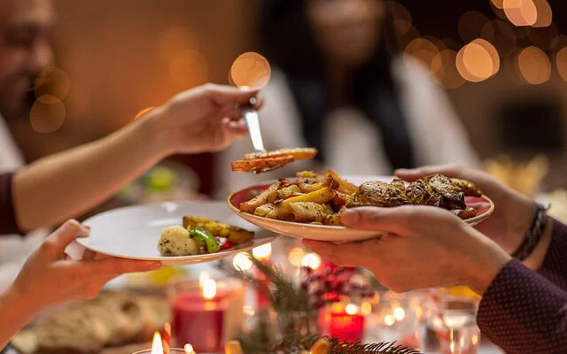 In einer Nahaufnahme ist eine festlich gedeckte Weihnachtstafel zu sehen. Kerzen brennen, im Ofen gebackene Gemüsestücke werden auf einen Teller gegeben.