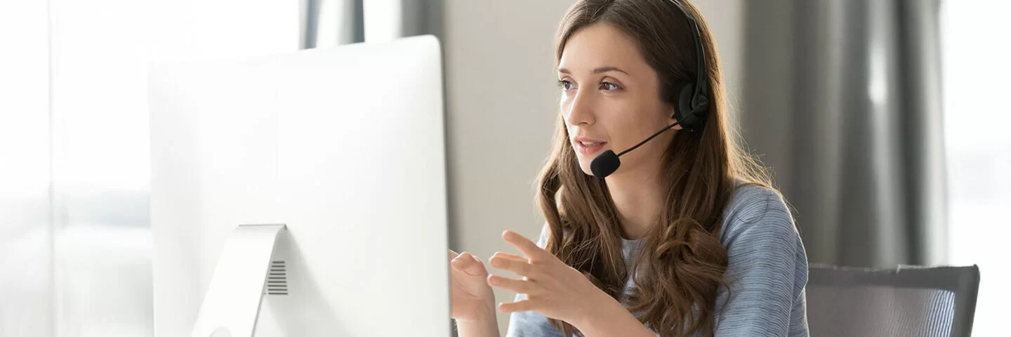 Eine junge Frau im drahtlosen Headset, spricht in ihr Mikrofon und blickt auf den Computerbildschirm.