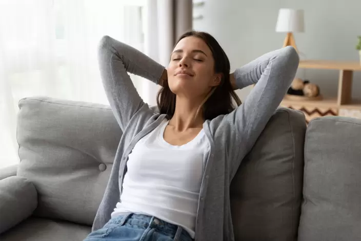 Eine junge Frau sitzt auf dem Sofa und macht mit hinter dem Kopf verschränkten Armen eine Pause.