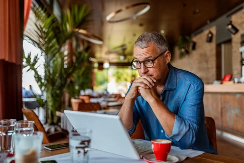 Ein Mann sitzt in einem gemütlichen Café. Auf dem Tisch vor ihm steht ein Laptop sowie weiteres Arbeitsmaterial. Er wirkt nachdenklich und angespannt.