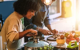 Eine Frau und ein Mann kochen gemeinsam in der Küche mit gesunden, frischen Zutaten, um einer Fettleber vorzubeugen.