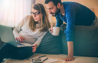 Eine Frau sitzt auf einer Couch. Hinter ihr steht ein Mann. Beide schauen auf einen Laptop, den die Frau auf ihrem Schoß hat.