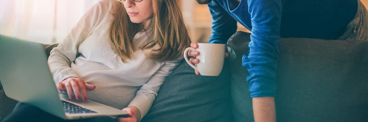 Eine Frau sitzt auf einer Couch. Hinter ihr steht ein Mann. Beide schauen auf einen Laptop, den die Frau auf ihrem Schoß hat.