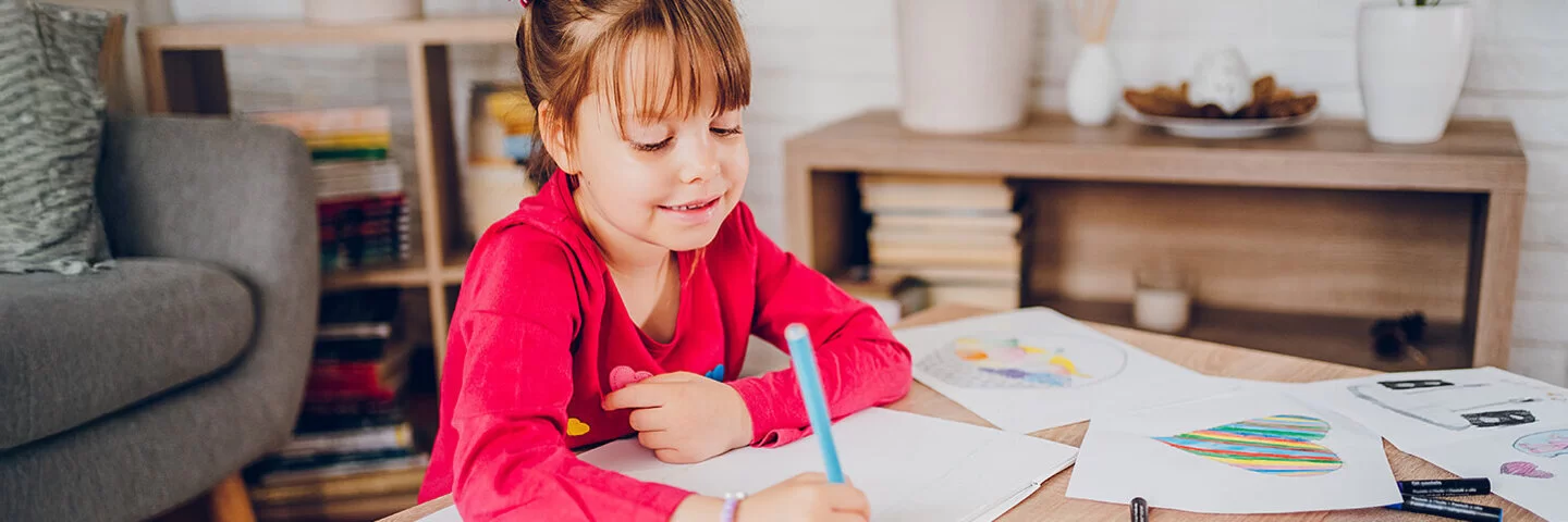 Ein kleines Mädchen sitzt zuhause an einem Tisch. Vor ihr liegen ein leeres weißes Blatt Papier und viele bunte Stifte. Am Rande des Tisches sind Blätter mit bunten Zeichnungen zu sehen. Das Mädchen hält in der rechten Hand einen blauen Stift und beginnt zu malen.