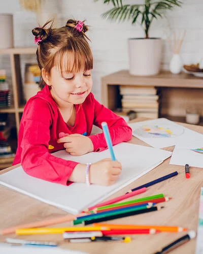 Ein kleines Mädchen sitzt zuhause an einem Tisch. Vor ihr liegen ein leeres weißes Blatt Papier und viele bunte Stifte. Am Rande des Tisches sind Blätter mit bunten Zeichnungen zu sehen. Das Mädchen hält in der rechten Hand einen blauen Stift und beginnt zu malen.