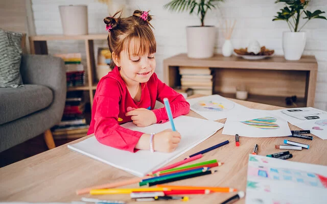 Ein kleines Mädchen sitzt zuhause an einem Tisch. Vor ihr liegen ein leeres weißes Blatt Papier und viele bunte Stifte. Am Rande des Tisches sind Blätter mit bunten Zeichnungen zu sehen. Das Mädchen hält in der rechten Hand einen blauen Stift und beginnt zu malen.