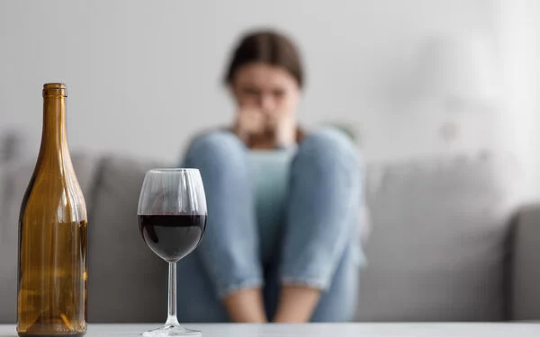 Eine Frau sitzt zusammengekauert auf einem Sofa. Vor ihr stehen ein gefülltes Weinglas und eine leere Flasche. 