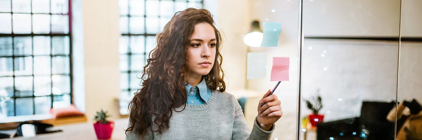 Eine junge Frau mit langen braunen Haaren steht allein in einem Konferenzraum und schaut konzentriert auf einige Haftnotizen, die an die Glaswand geklebt sind. Auf eine davon tippt sie mit einem Kugelschreiber. In der anderen Hand hält sie eine Kaffeetasse.