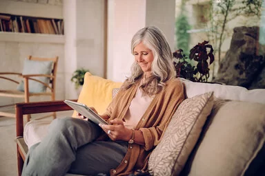 Eine ältere Frau mit langen grauen Haaren sitzt entspannt auf einem gemütlichen Sofa in einem hellen Wohnzimmer. Sie trägt einen beigefarbenen Cardigan und hält ein Tablet in den Händen, auf das sie lächelnd schaut. Im Hintergrund sind Pflanzen, Regale und ein Sessel zu sehen, was dem Raum eine einladende Atmosphäre verleiht.